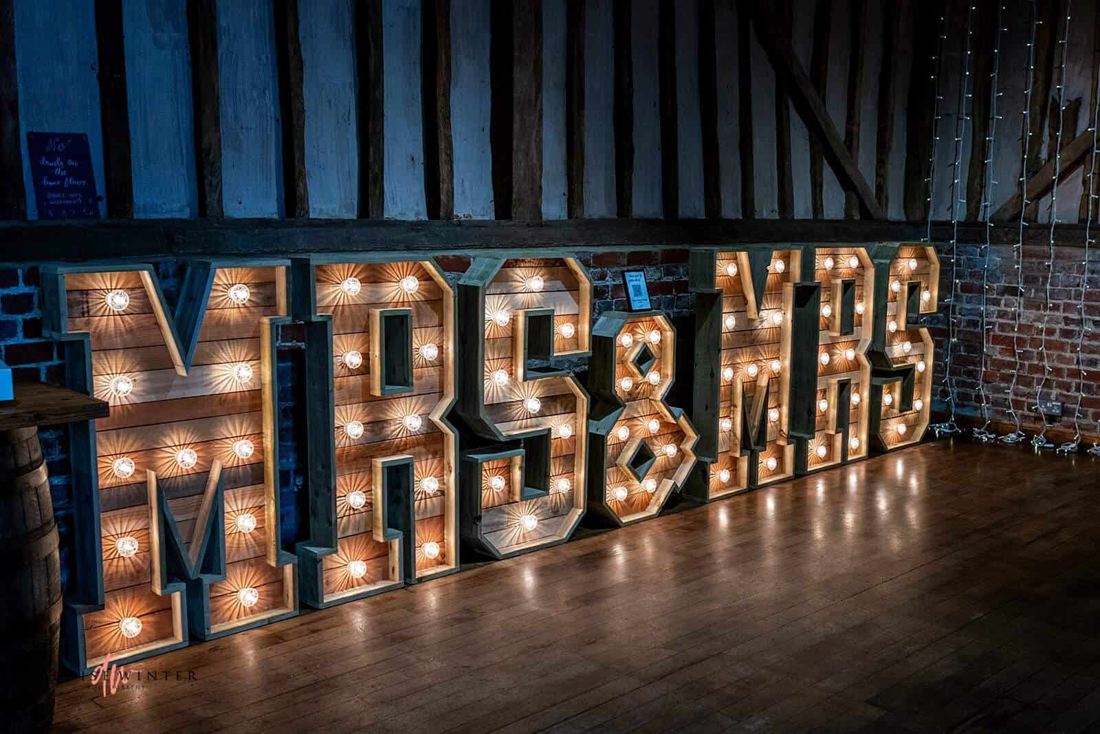 The image features a decorative display of illuminated letters spelling out "MRS & MRS." The letters are large and adorned with bright bulbs, creating a warm and inviting glow. Set against a rustic backdrop of wooden beams and brick walls, this charming piece adds a romantic touch to the venue, making it a perfect focal point for a wedding celebration. The floor is wooden, complementing the overall rustic aesthetic of the space.