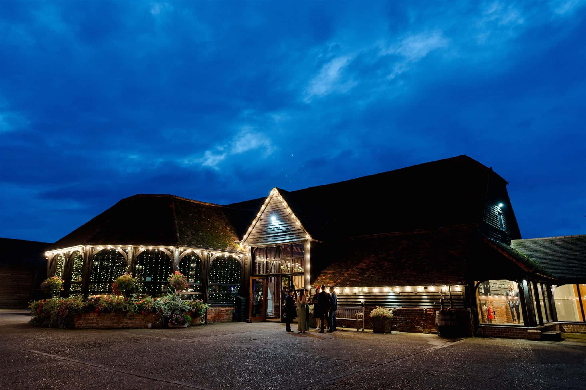 The image depicts the Great Barn at Lillibrooke Manor, beautifully illuminated with string lights against a twilight sky. The barn features a large entrance with glass windows and a spacious facade. A few people are seen approaching the entrance, creating a warm and inviting atmosphere.