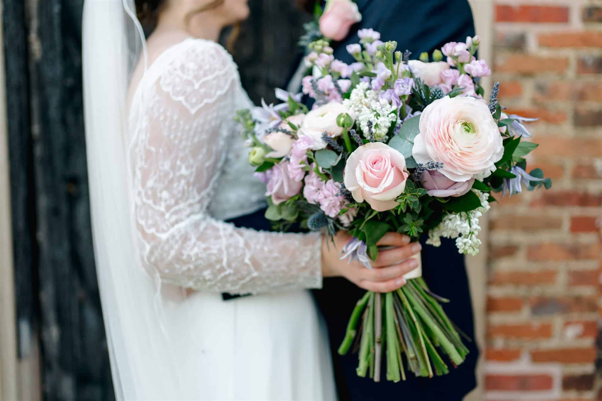 A bride and groom share a tender moment at a wedding venue in Berkshire, with the bride holding a stunning bouquet filled with soft pink roses and delicate wildflowers. She is dressed in a beautifully detailed gown with long lace sleeves, while the groom stands beside her, dressed elegantly. The background features rustic brick, adding charm and warmth to the romantic scene. The overall atmosphere captures the joy and love of their special day.