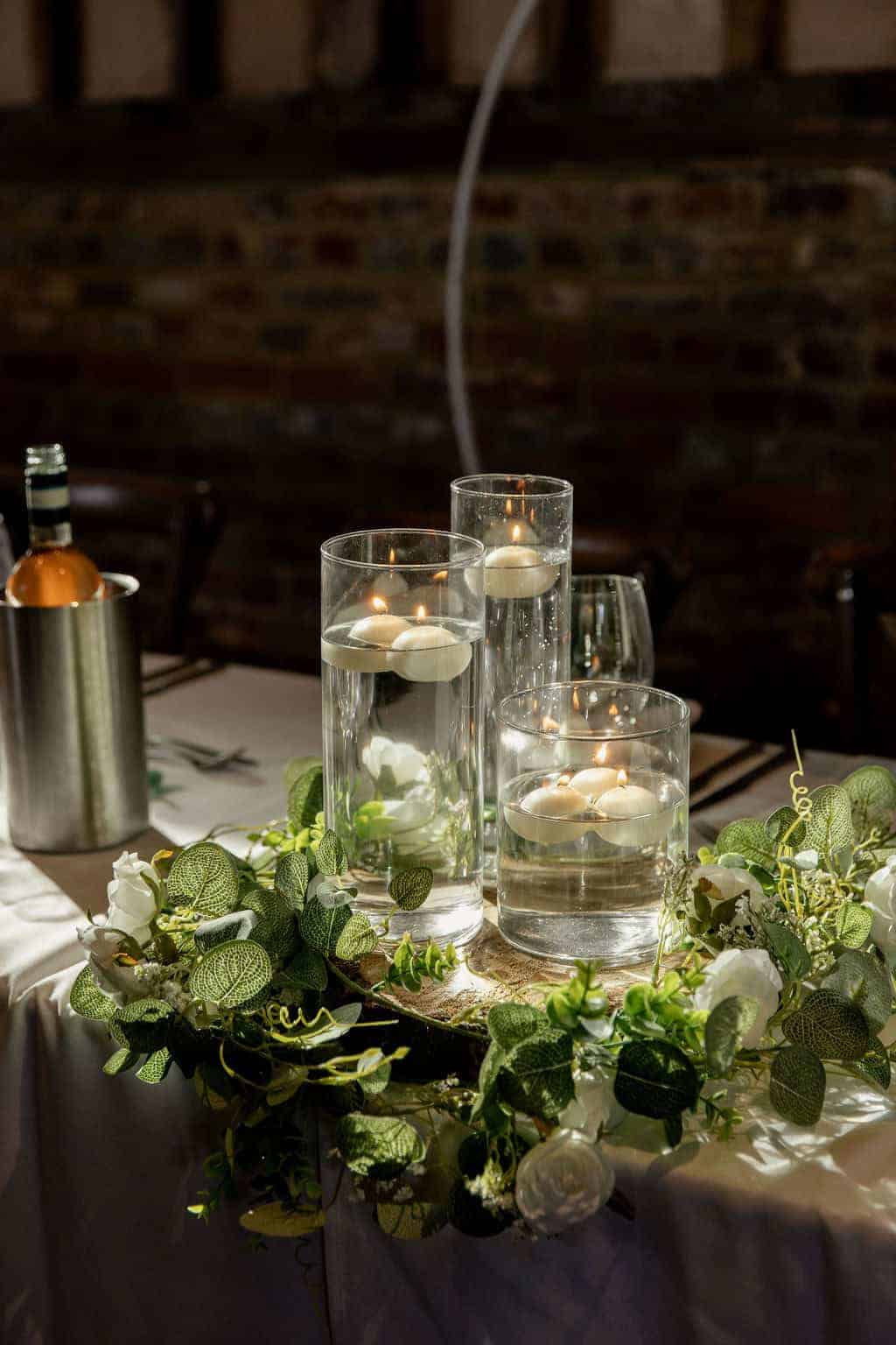The image features an elegant table centerpiece consisting of three tall glass vases filled with water and floating candles. Surrounding the vases is a lush arrangement of greenery and white flowers, enhancing the romantic ambiance. A silver ice bucket holding a bottle is positioned beside the centerpiece, contributing to the sophisticated table setting in a rustic venue with a warm