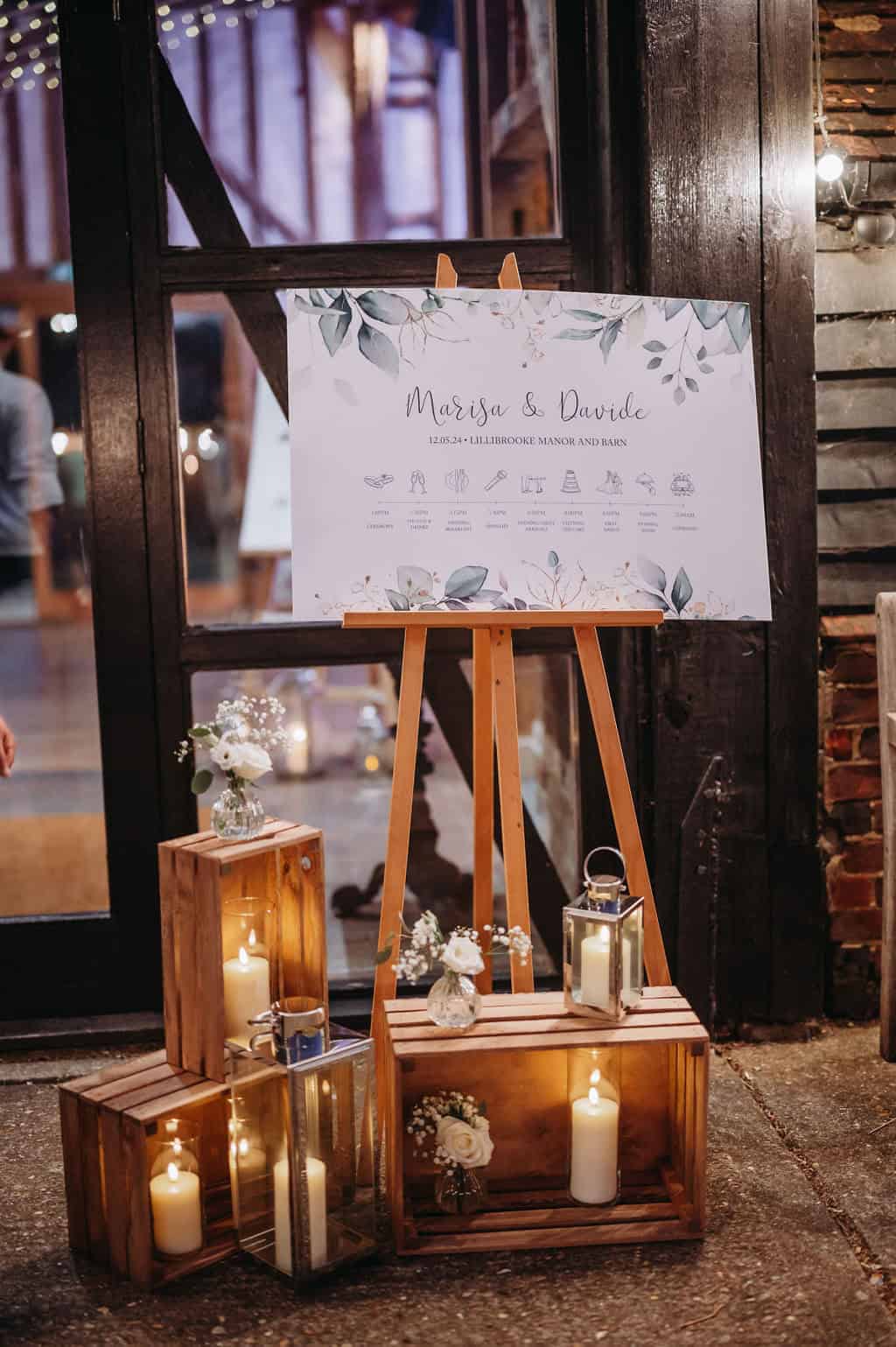 The image shows a welcome display outside the Great Barn at Lillibrooke. An easel holds a beautifully designed sign featuring the names "Margo & Daniel," along with event details, adorned with greenery. Surrounding the easel are wooden crates and lit candles, creating a cozy and inviting atmosphere for guests. The setting suggests a rustic charm typical of a barn venue.