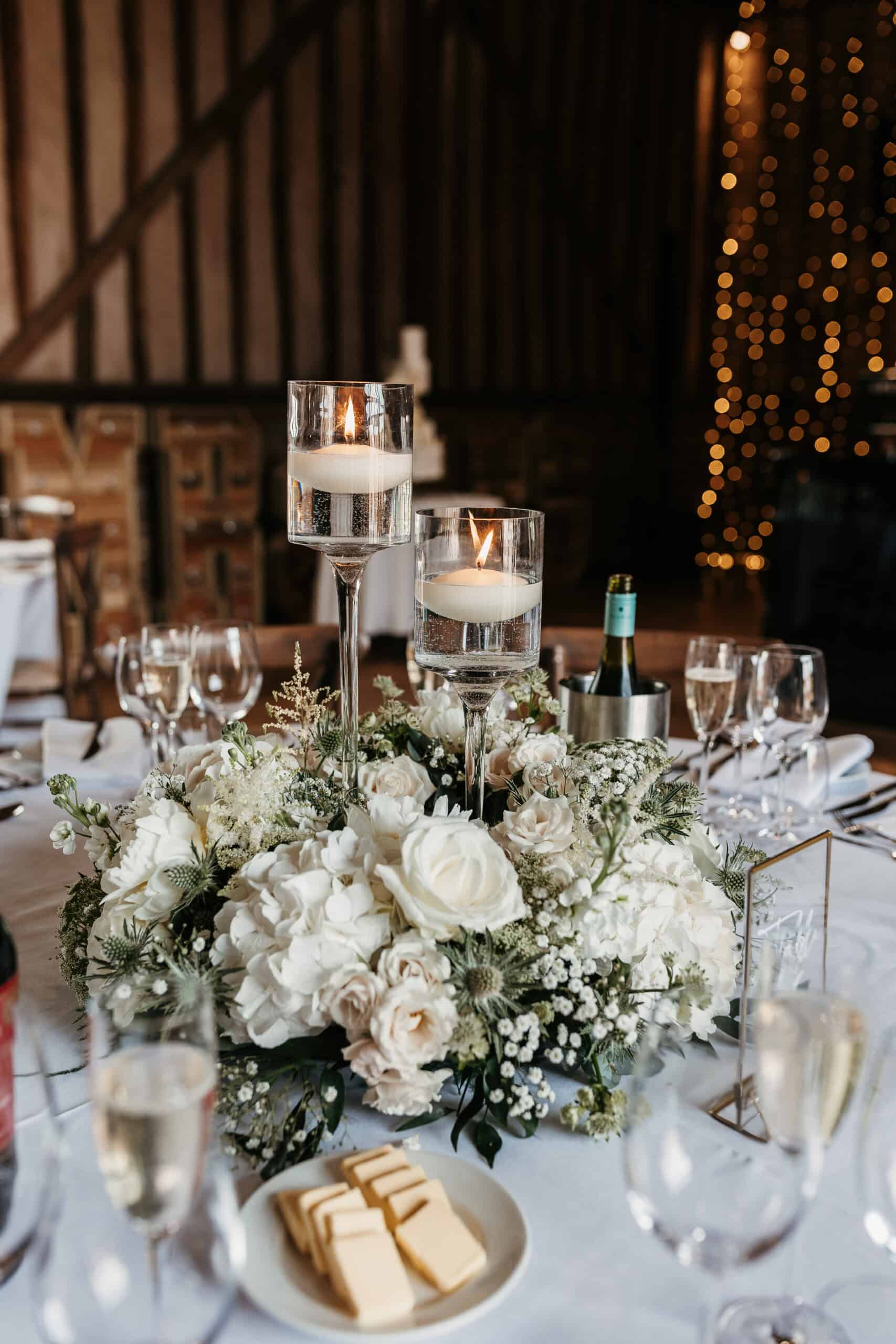 The image features an elegant table centerpiece consisting of three tall glass vases filled with water and floating candles. Surrounding the vases is a lush arrangement of greenery and white flowers, enhancing the romantic ambiance. A silver ice bucket holding a bottle is positioned beside the centerpiece, contributing to the sophisticated table setting in a rustic venue with a warm