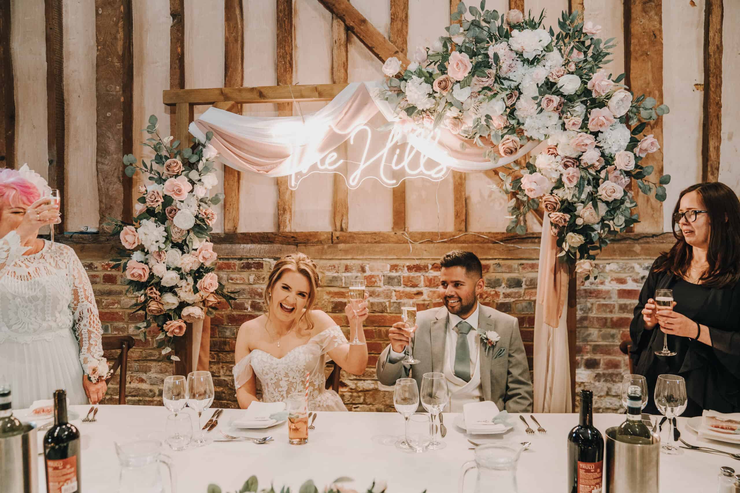The image shows a wedding reception scene featuring a bride and groom seated at a beautifully decorated table. The bride, wearing a stunning white gown, is smiling and raising a glass, while the groom, dressed in a light grey suit, is beside her. Floral arrangements with pink and white flowers frame the backdrop, along with a neon sign that reads "The Mills" Two additional guests, one in a light-colored dress and another in a black dress, are also present, creating a festive atmosphere in a rustic venue with wooden beams.