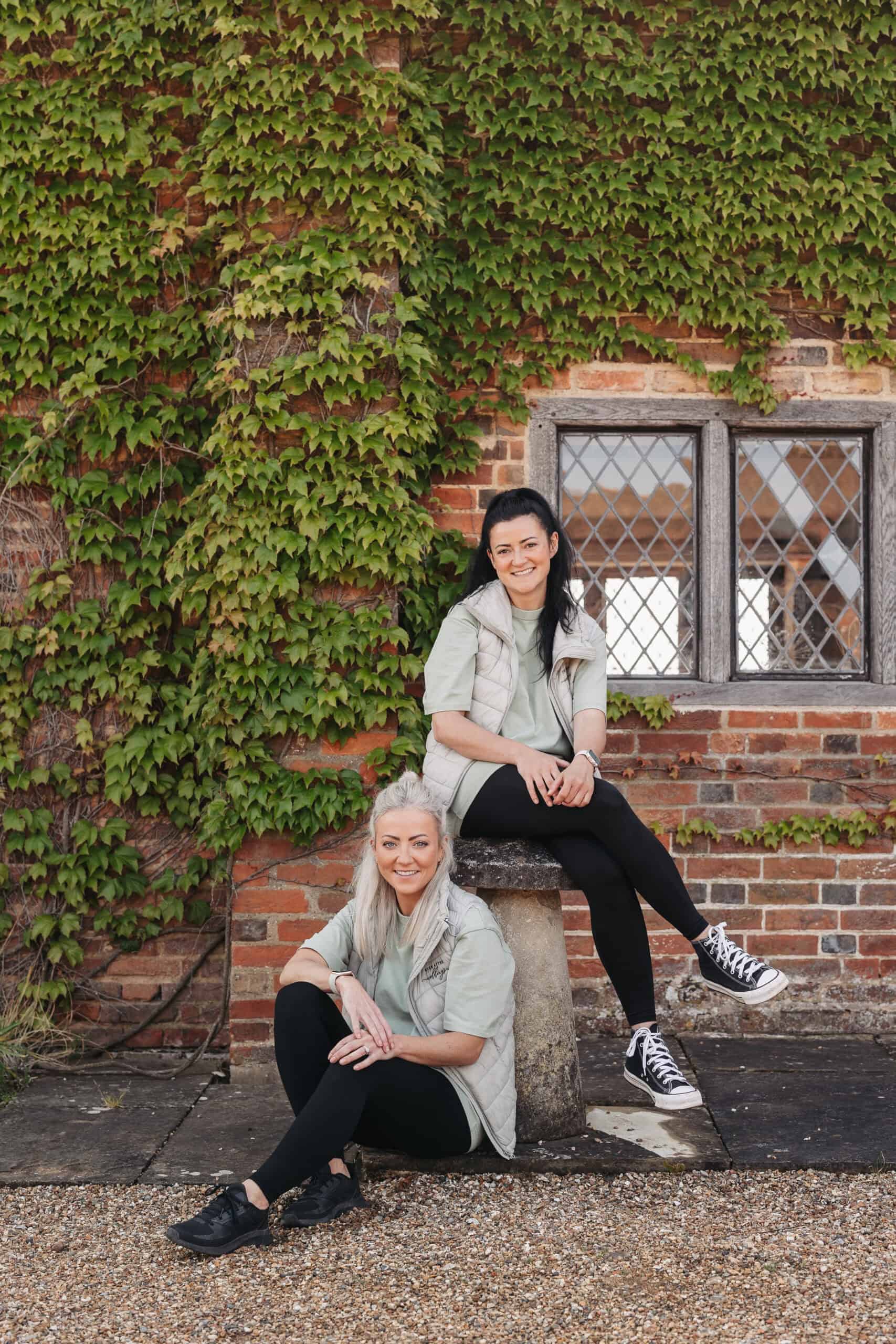 Two florists from Ever After Weddings sitting against a brick wall adorned with ivy, smiling and showcasing their creative floral design expertise.