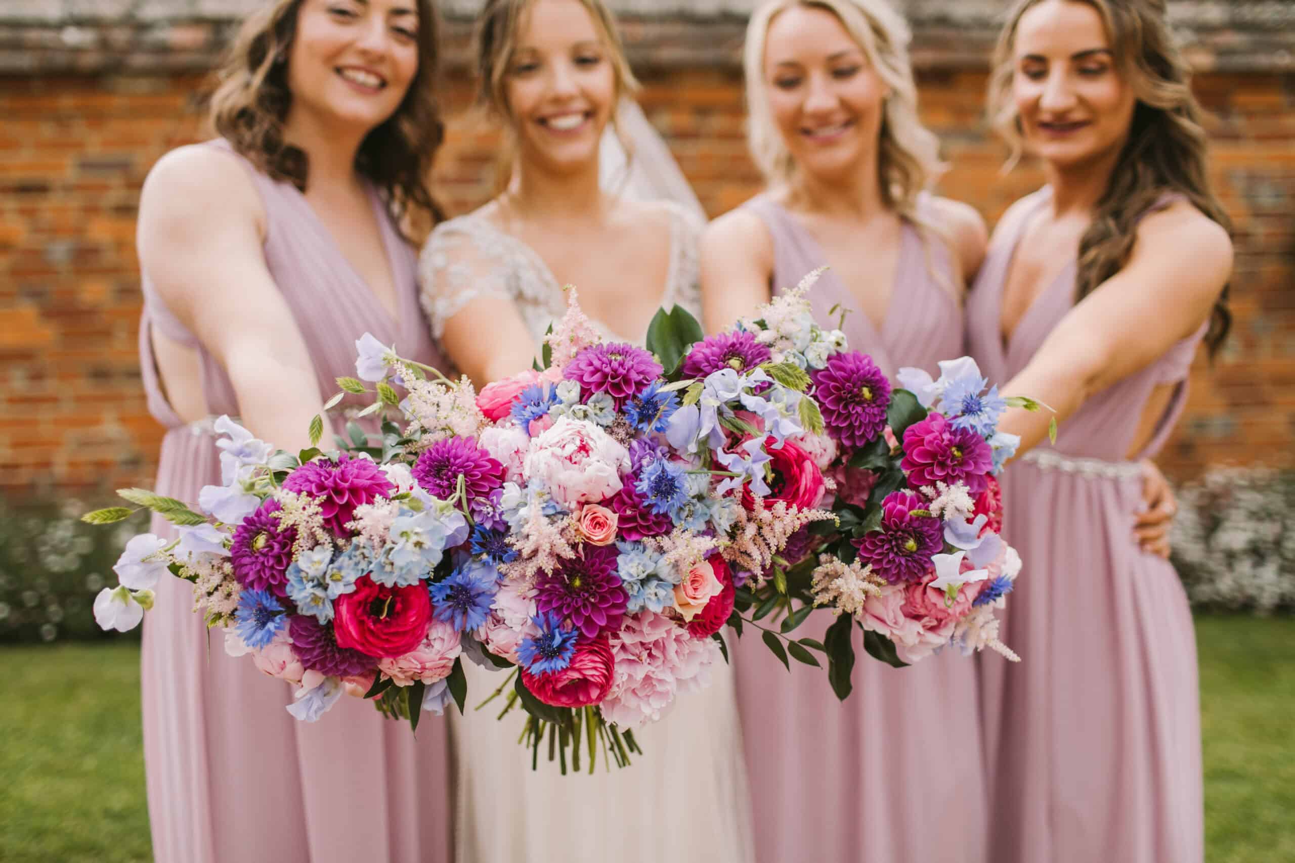 A bride stands in the center, surrounded by three bridesmaids, each wearing flowing lavender dresses. They are all smiling and holding vibrant bouquets filled with a mix of colorful flowers, including pinks, purples, and blues. The backdrop features a rustic brick wall and hints of greenery, adding to the joyful and celebratory atmosphere of the moment.