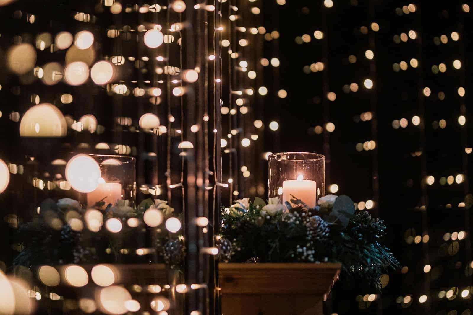 The image features a close-up view of a beautifully decorated table setting with soft, glowing lights in the background. In the foreground, a clear glass candle holder contains a lit candle, providing a warm and inviting glow. Surrounding the candle are decorative elements, including greenery and floral arrangements, that enhance the romantic atmosphere. The overall effect is enchanting, creating a cozy and elegant ambiance, perfect for a special event or celebration.