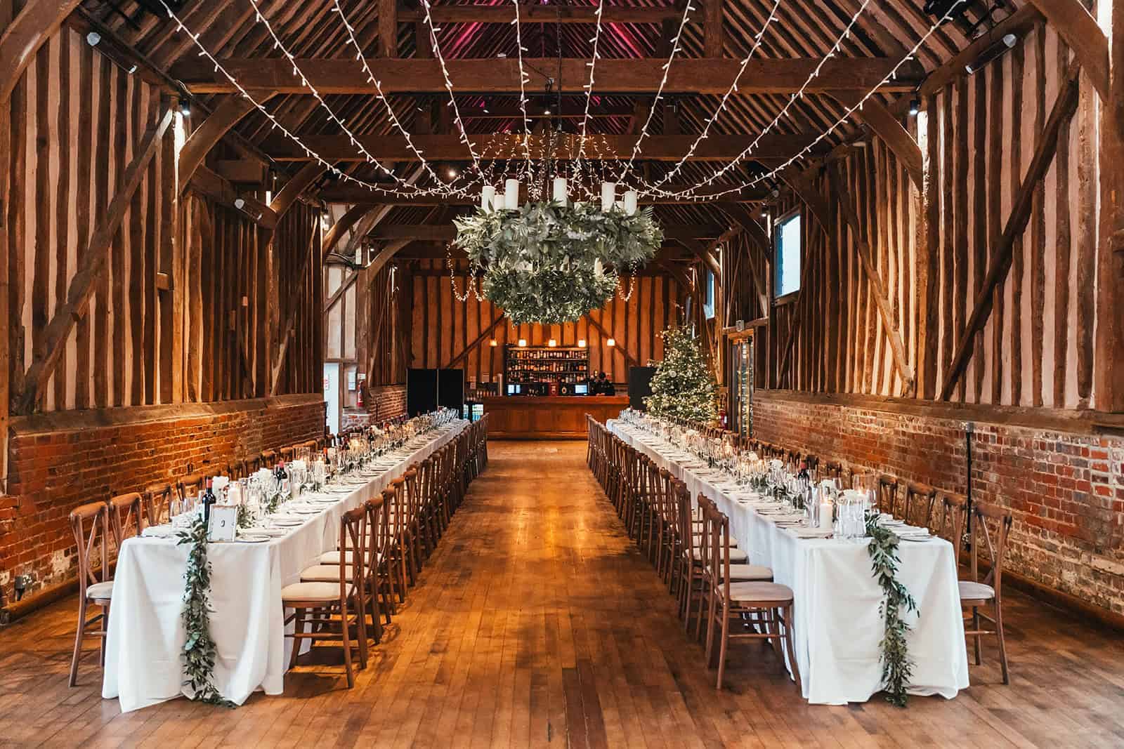 The image depicts the interior of The Great Barn at Lillibrooke, showcasing a stunning event space with a long dining setup. Two rows of elegantly dressed tables with white tablecloths are arranged in the center, flanked by wooden chairs. Above, a large greenery chandelier hangs from the wooden beams, while string lights add a warm glow to the rustic setting. A beautifully decorated Christmas tree stands in the corner, enhancing the festive atmosphere.