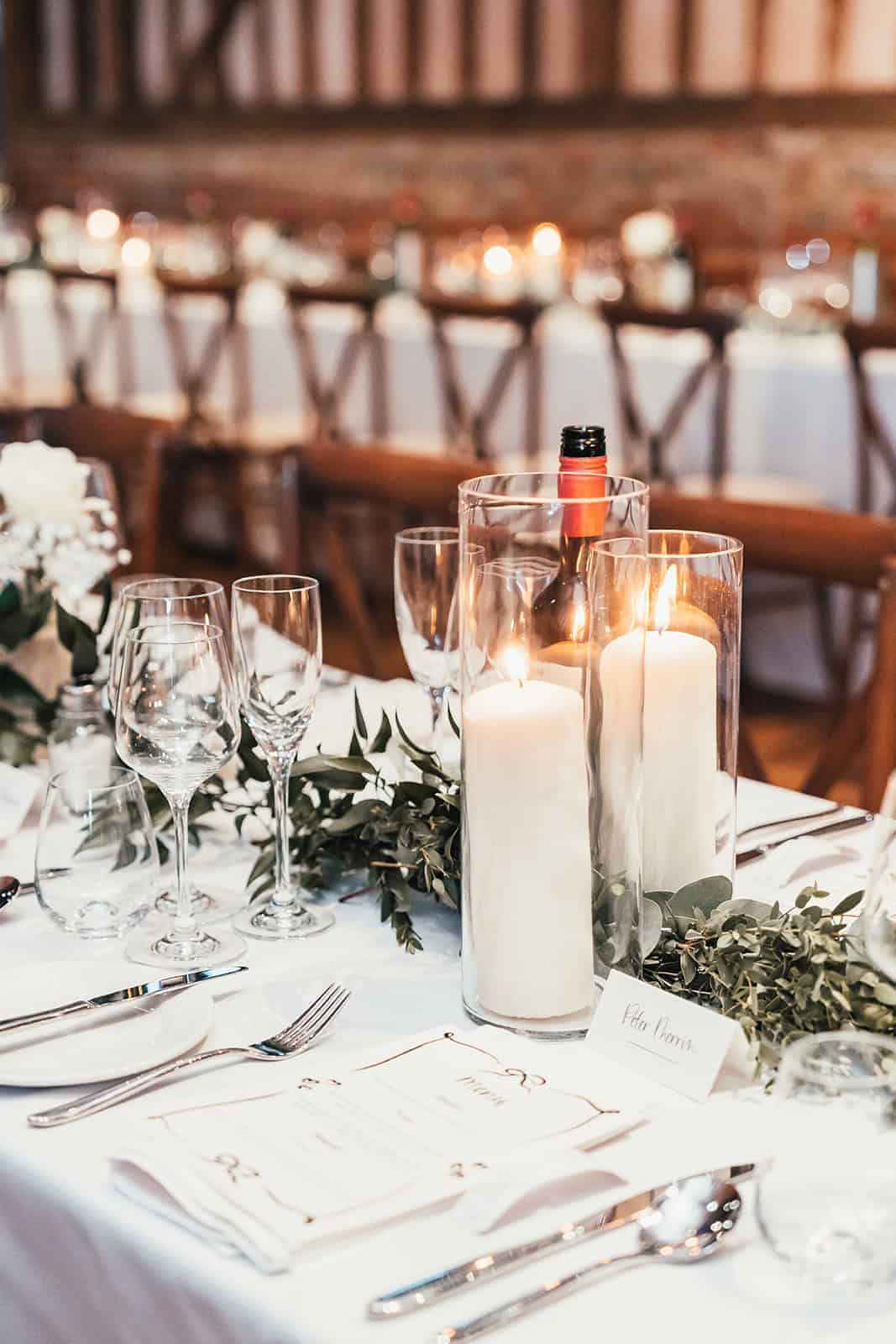 A beautifully set dining table adorned with glassware, delicate white flowers, and a tall candle in a glass holder, creating an elegant atmosphere for a wedding.