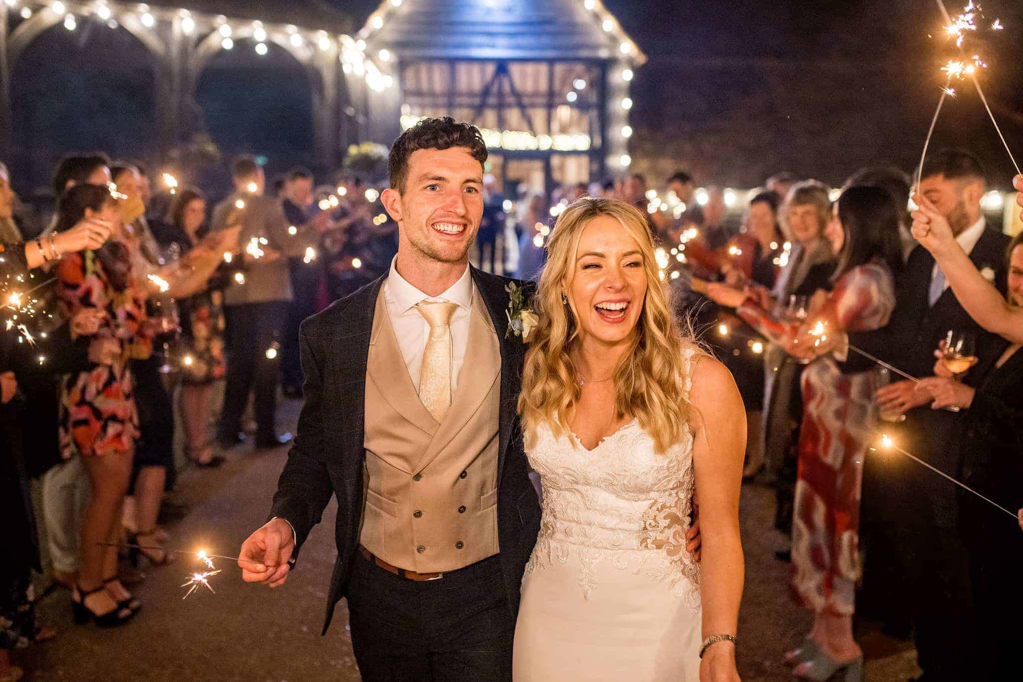 The image captures a joyful moment of a newlywed couple exiting their wedding celebration. The bride, wearing a stunning white gown, and the groom, dressed in a black suit with a beige vest, are smiling and holding hands as they walk through a crowd of guests. Surrounding them are friends and family holding sparklers, creating a magical and festive atmosphere. The backdrop features twinkling fairy lights and a barn-like structure, enhancing the celebratory mood of the evening. The scene radiates happiness and love, marking a memorable moment in the couple's life.