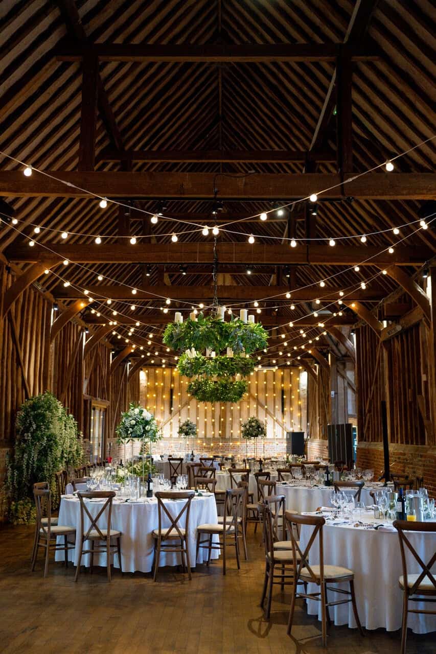 The image showcases the interior of the Great Barn at Lillibrooke Manor, featuring a spacious barn setting adorned with wooden beams and rustic charm. The room is illuminated by string lights hanging from the ceiling, creating a warm and inviting atmosphere. Round tables are elegantly set with white tablecloths and centerpieces, while a large greenery chandelier hangs above the main area, enhancing the decor. The overall ambiance is perfect for events, combining elegance with a natural, rustic feel.
