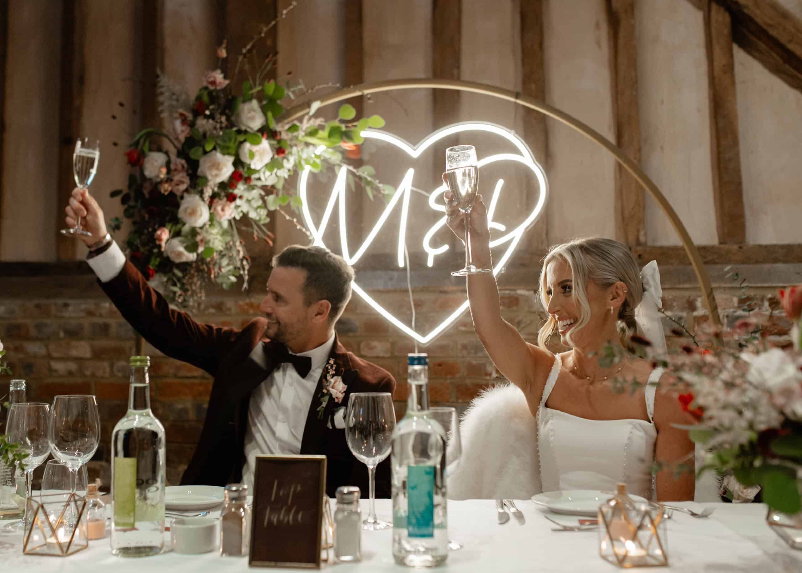 The image captures a joyful moment at a wedding reception featuring a couple toasting with glasses. The groom, dressed in a black tuxedo, and the bride, in a white gown, are seated at a beautifully decorated table adorned with floral arrangements. Behind them, a neon sign in the shape of a heart displays the initials "M&L." The rustic venue has wooden beams, adding to the celebratory atmosphere.