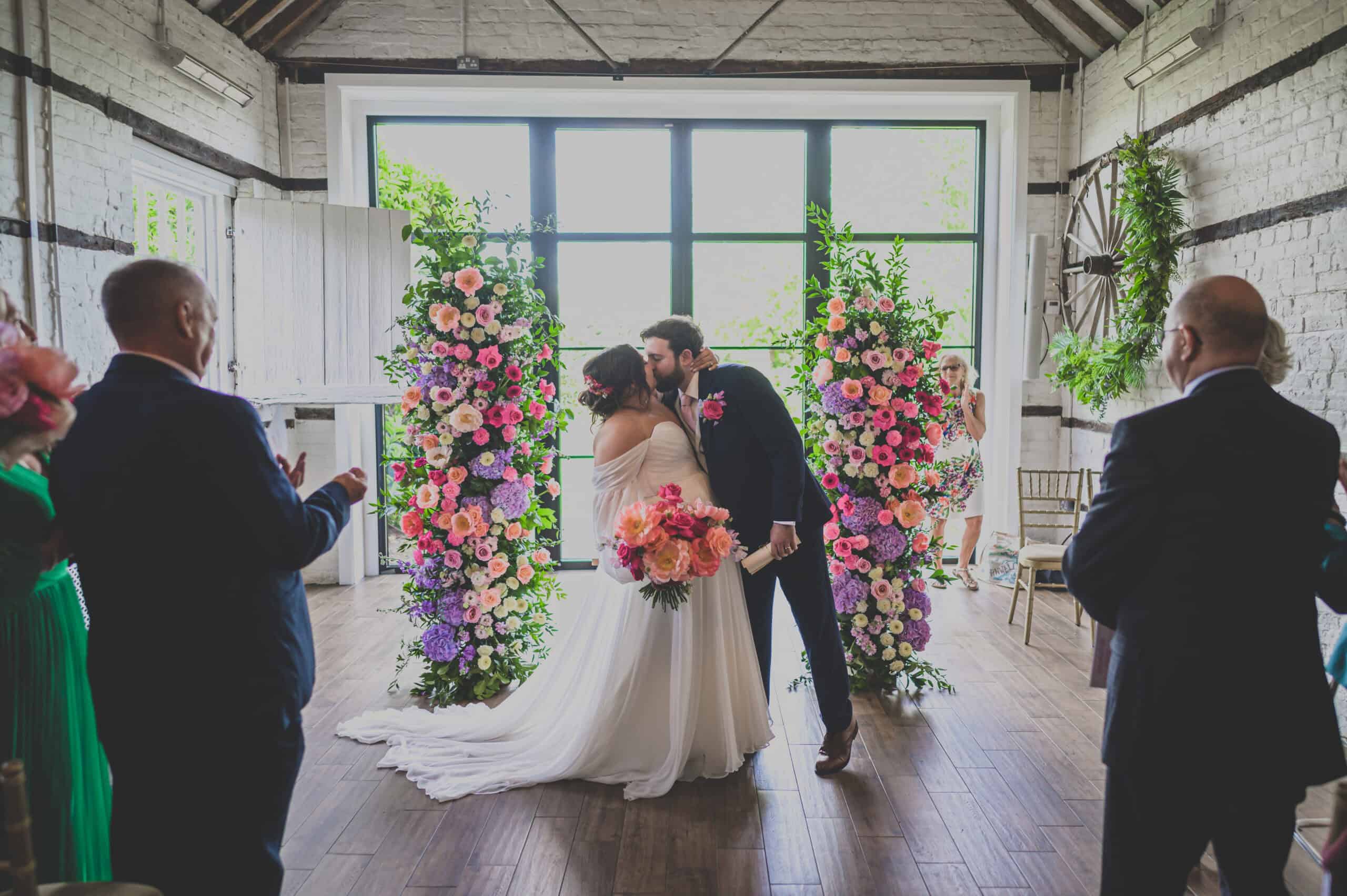 A just-married couple sharing a kiss in the small barn at Lillibrooke Manor, surrounded by vibrant floral arrangements and applauding guests