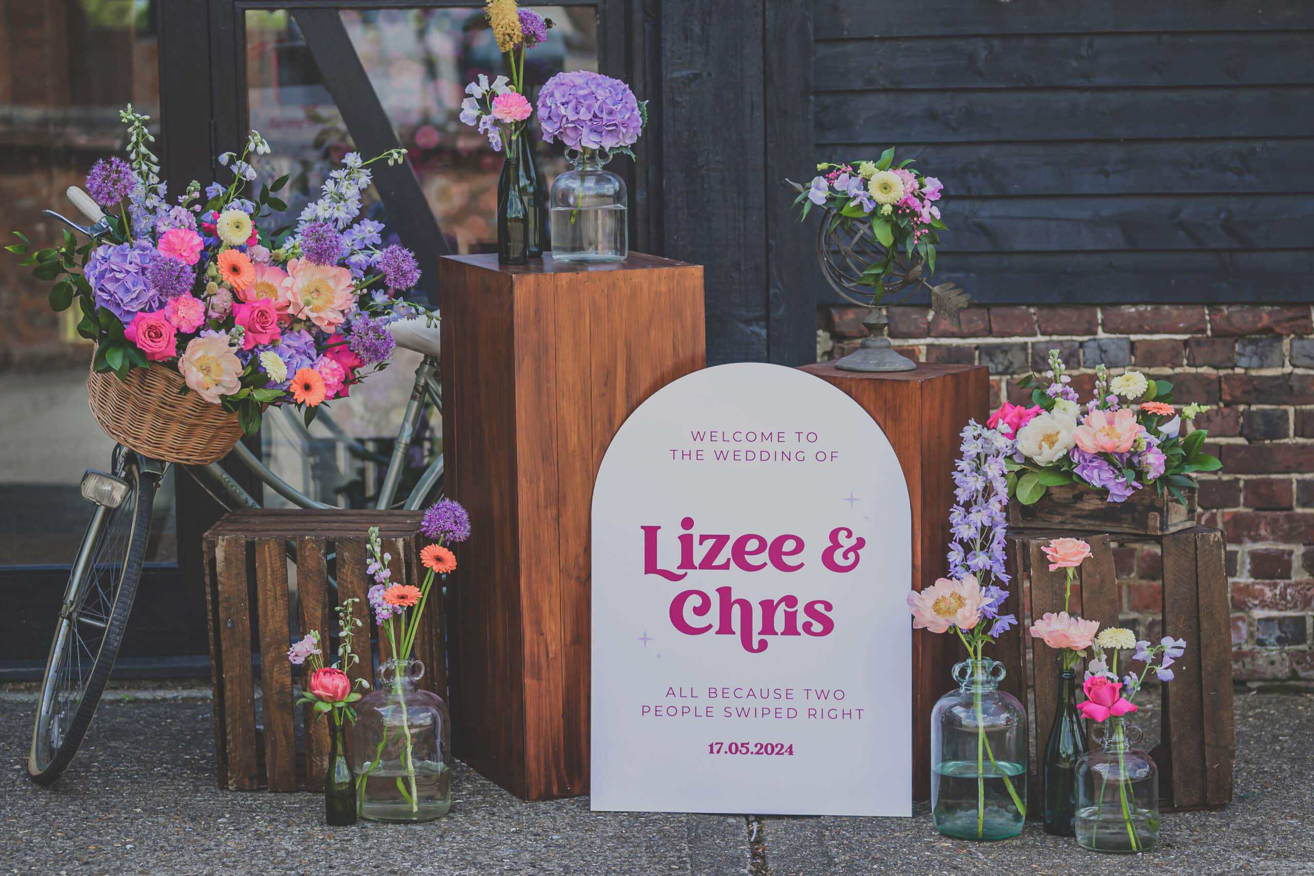 A beautifully arranged welcome display for Lizze and Chus's wedding at Lillibrooke Manor just outside the Great Barn, featuring vibrant flowers in various containers alongside a sign announcing the event.